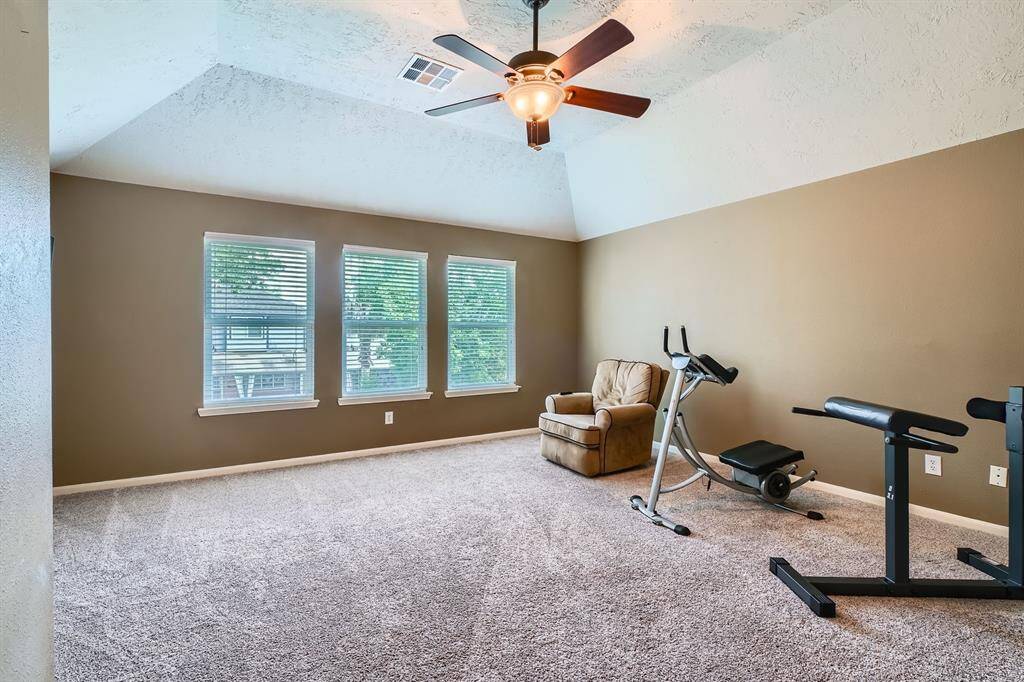 This spacious, carpeted room features a vaulted ceiling with a fan, three large windows offering natural light, and room for exercise equipment or home furnishings.