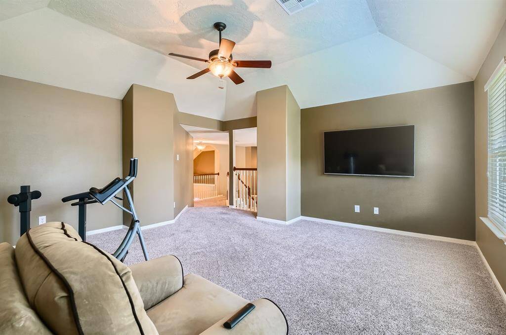 This spacious room features a plush carpet, neutral walls, and ample natural light from large windows. It's equipped with a ceiling fan, wall-mounted TV (not incl), and space for exercise equipment, offering a versatile area for relaxation or workouts.