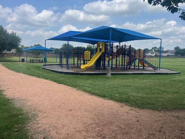 This photo shows a well-maintained playground with colorful equipment, including slides and climbing structures, covered by blue shade canopies. It's situated in a grassy area with a walking path, ideal for families with children.