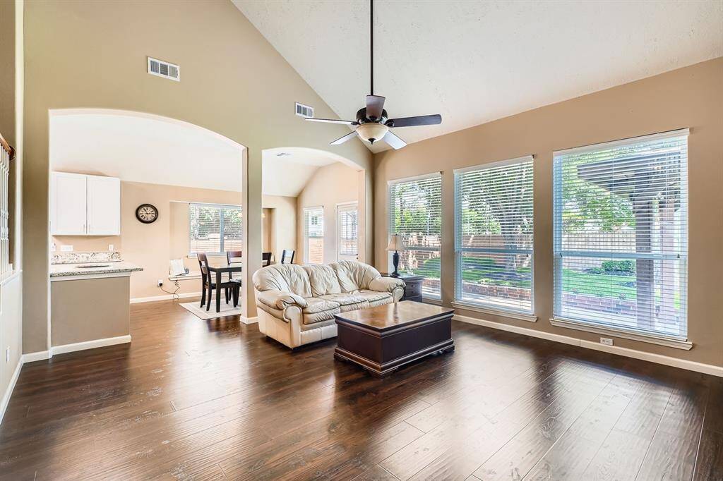This bright, open concept living area features high ceilings and large double pain windows overlooking a lush backyard. The space flows seamlessly into a dining area and kitchen, enhancing the home's airy feel.
