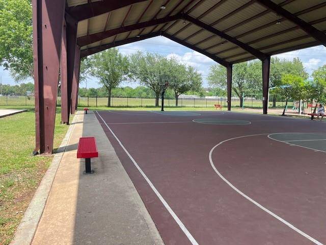This photo features a covered outdoor basketball court with a metal roof, providing shade and protection from the elements. The area is surrounded by greenery, including trees and a grassy field, offering a pleasant and spacious environment for recreational activities.