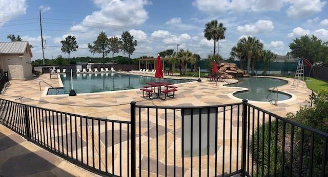 Spacious outdoor community pool area with a fenced perimeter. It features multiple pools, lounge chairs, red umbrellas, and a rock waterfall. The setting is surrounded by greenery and palm trees, providing a resort-like atmosphere.