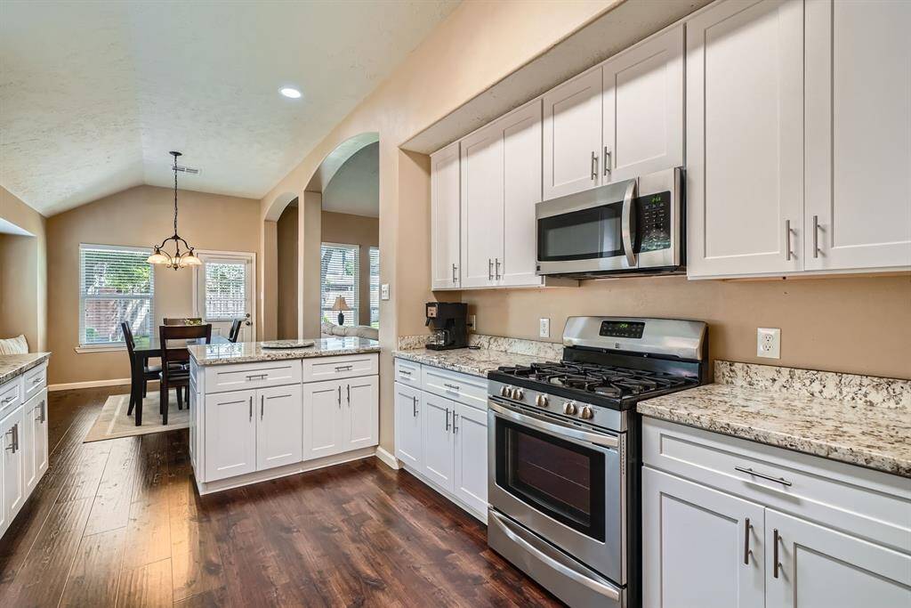 This photo showcases a modern kitchen with sleek white cabinetry, granite countertops, and stainless steel appliances. The open layout leads to a bright dining area with large windows and wood flooring, creating a warm and inviting space.