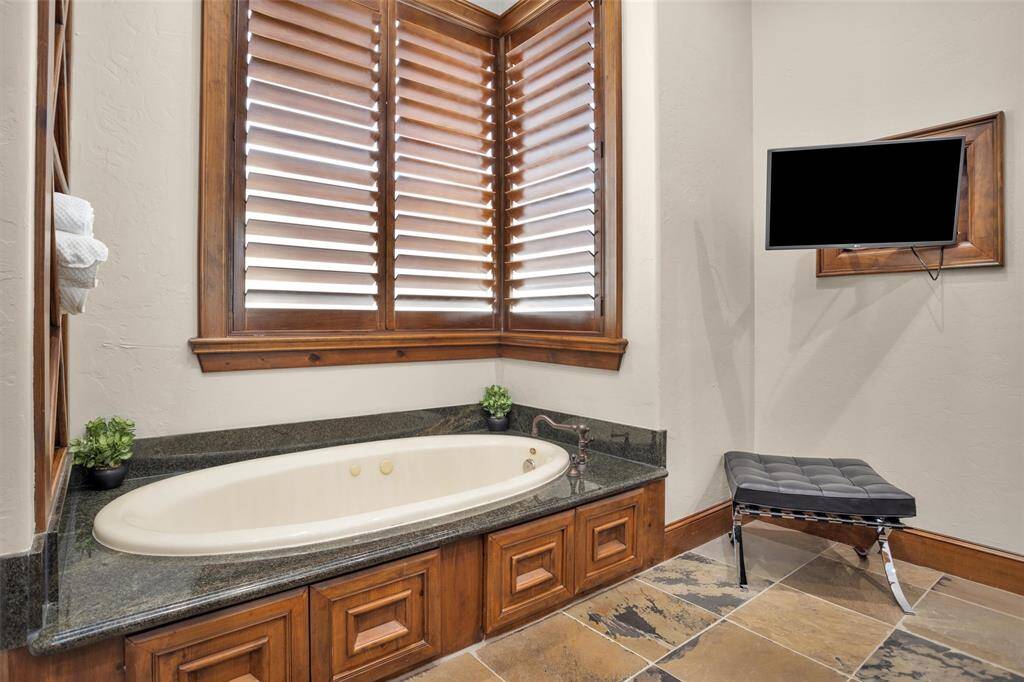 The jetted soaking tub is surrounded by granite, with a built-in TV and bench nearby for a spa-like escape right at home.