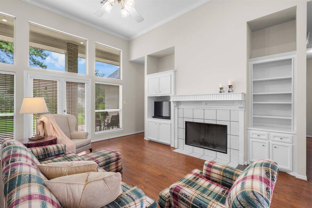 This spacious living room features high ceilings, ceiling fan, abundant natural light from large windows, and a cozy gas log fireplace with built-in shelving. The open layout is complemented by warm wooden flooring and a neutral color palette, perfect for comfortable living and entertaining.