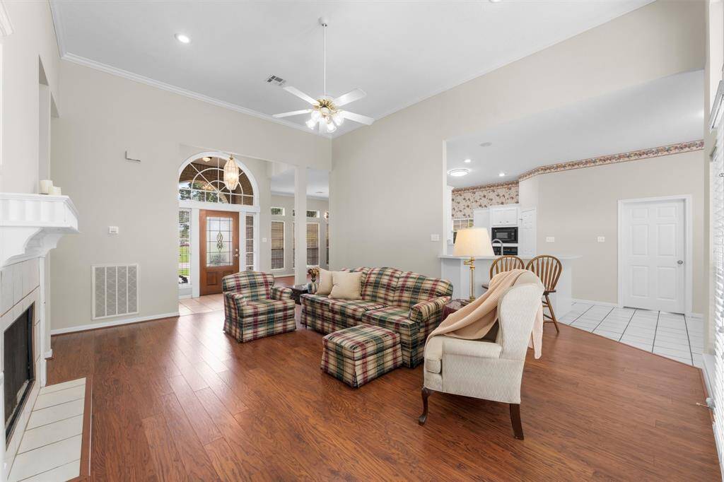 View from the Primary Suite hallway into the spacious, freshly painted bright living area that features wood flooring, a cozy fireplace, and high ceilings. It opens to a classic kitchen with tile flooring. The large entryway, with a beautiful glass front door, enhances the open feel.