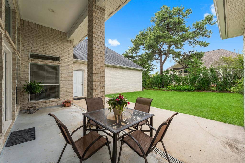 The rear covered back patio overlooks a well-maintained spacious lawn and is bordered by lush greenery, providing a serene outdoor space for relaxation or entertaining.