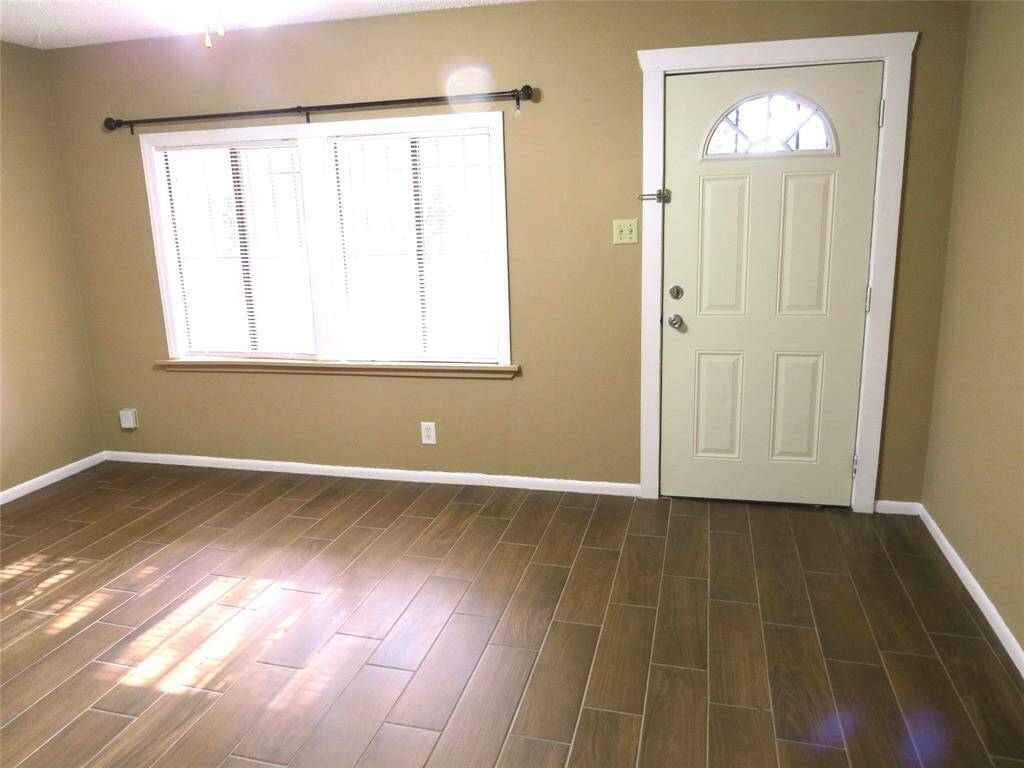 This room features wood-look tile flooring, neutral beige walls, a white-trimmed door with a decorative window, and a large window with blinds, offering ample natural light.
