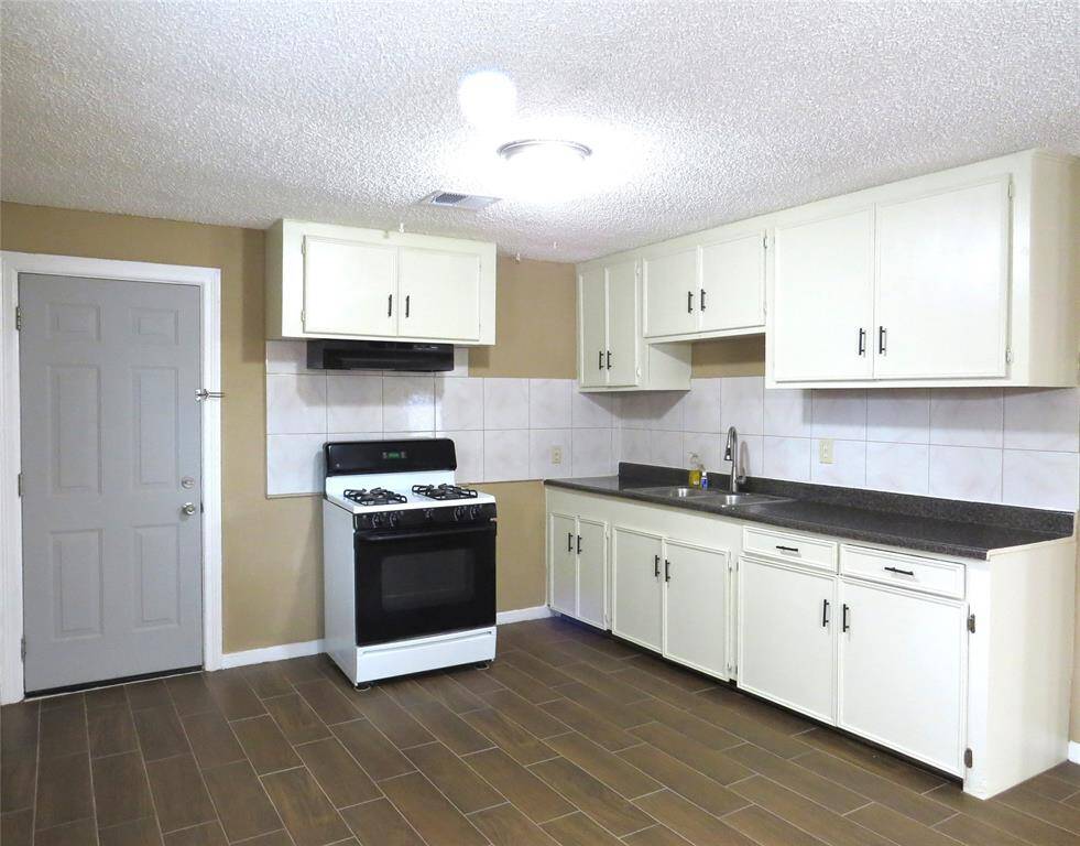 This kitchen features a clean and functional design with white cabinets, a gas stove, and tiled backsplash. It has dark countertops, a double sink, and wood-style flooring, offering a practical and inviting space.