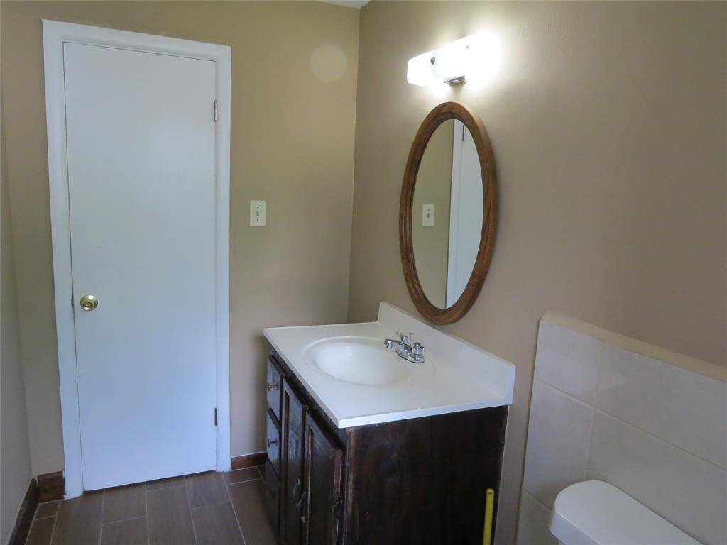 This is a compact bathroom with neutral-colored walls and floor tiles. It features a single sink vanity with a wood-framed mirror above and a well-lit overhead light. There's a white door and part of the toilet is visible.