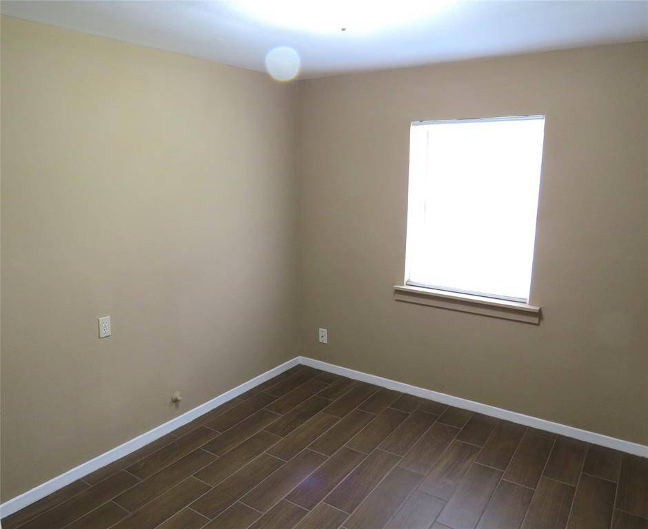 This is a small, unfurnished room with beige walls, a single window for natural light, and wood-look tile flooring.