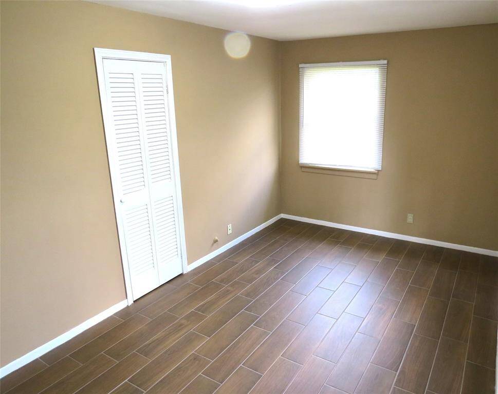 This photo shows a bright, empty room with light brown walls and wood-look tile flooring. It features a window with blinds and a white closet door.