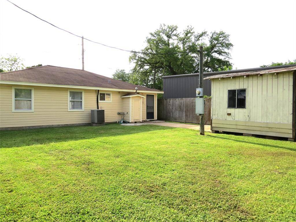 This photo shows a modest backyard with a well-maintained lawn, a small yellow house with a brown roof, and a separate shed or outbuilding. The space is open and bordered by a wooden fence, offering a private and cozy outdoor area.
