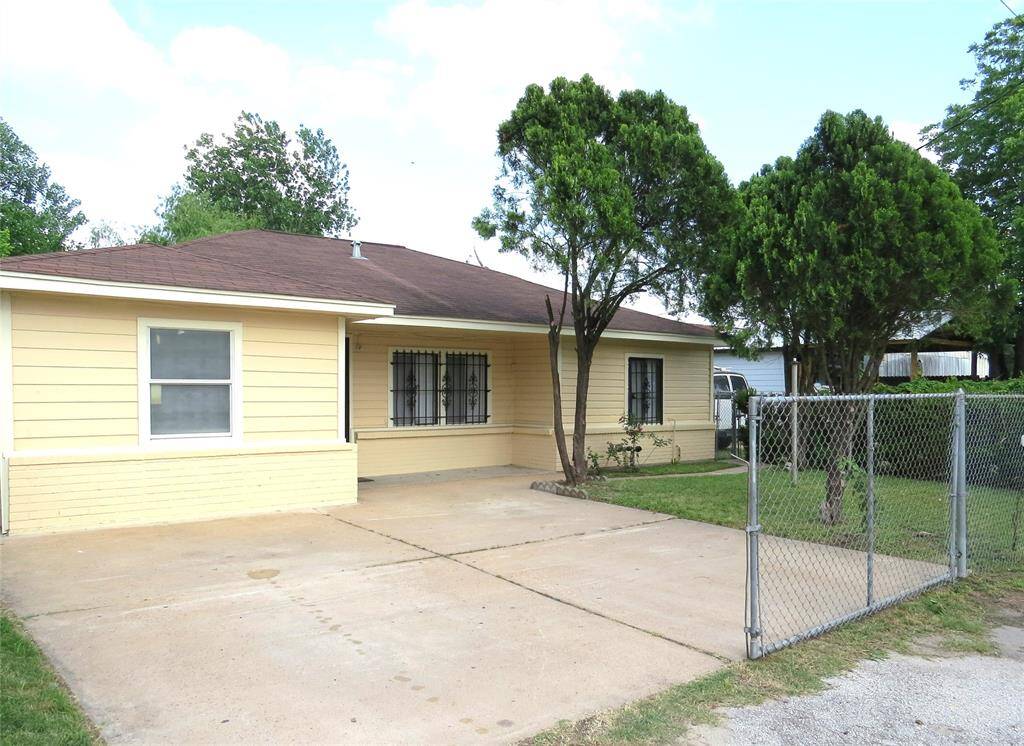 Charming yellow single-story home with a fenced driveway and gated entry. Features a neatly maintained yard with trees and a concrete parking area.
