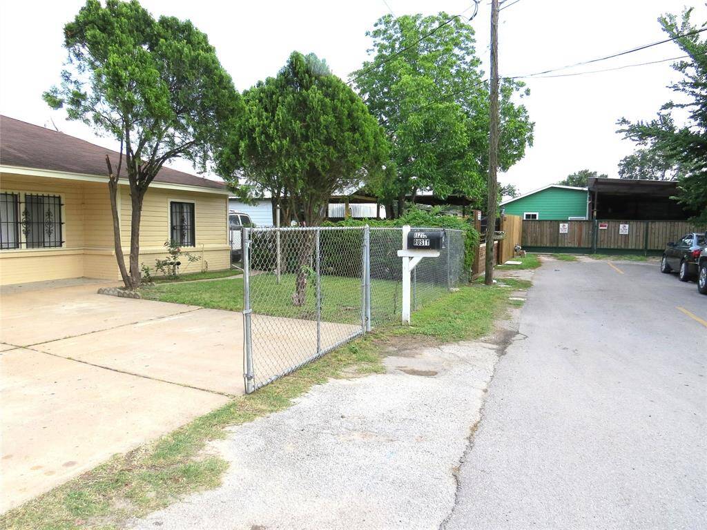 Charming home with a yellow exterior and secure window bars, featuring a gated driveway and a small front yard with mature trees. Located on a quiet street with a mailbox and nearby parking.