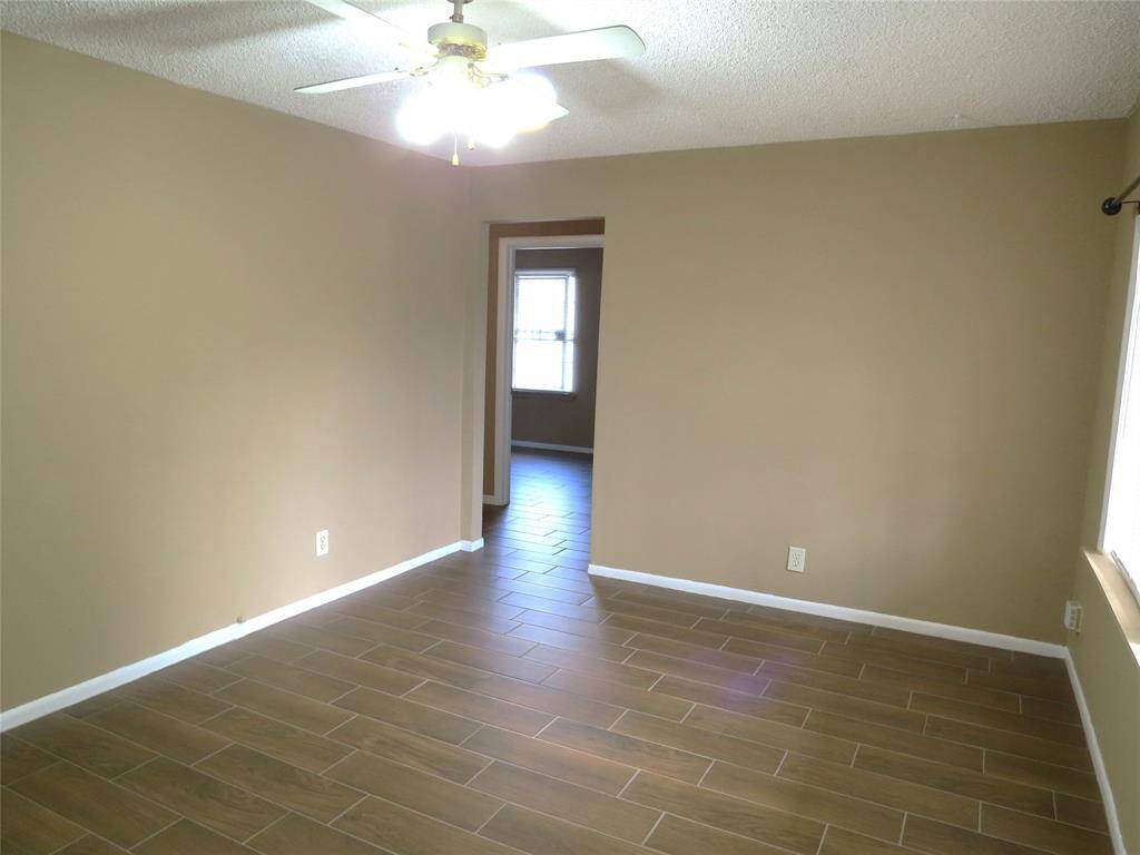 This room features neutral-colored walls, wood-look tile flooring, a ceiling fan, and ample natural light from a large window, creating a bright and welcoming space.