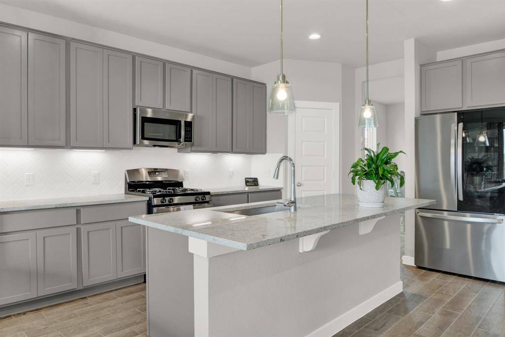 A modern kitchen featuring sleek gray cabinets, stainless steel appliances, and a spacious island with pendant lighting. The room includes a built-in microwave, gas stove, and a bright, clean backsplash, creating a stylish and functional space.