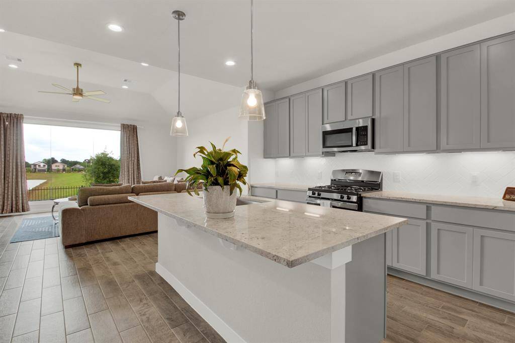 This modern kitchen features sleek gray cabinetry, stainless steel appliances, and a spacious island with pendant lighting. The open layout extends to a cozy living area with large windows offering a scenic outdoor view, perfect for entertaining.