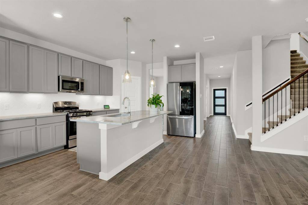 This modern kitchen features sleek gray cabinetry, stainless steel appliances, and a spacious island with pendant lighting. The open layout flows into a hallway with a stylish door and adjacent staircase, all highlighted by wood-look flooring. Perfect for entertaining and family living.
