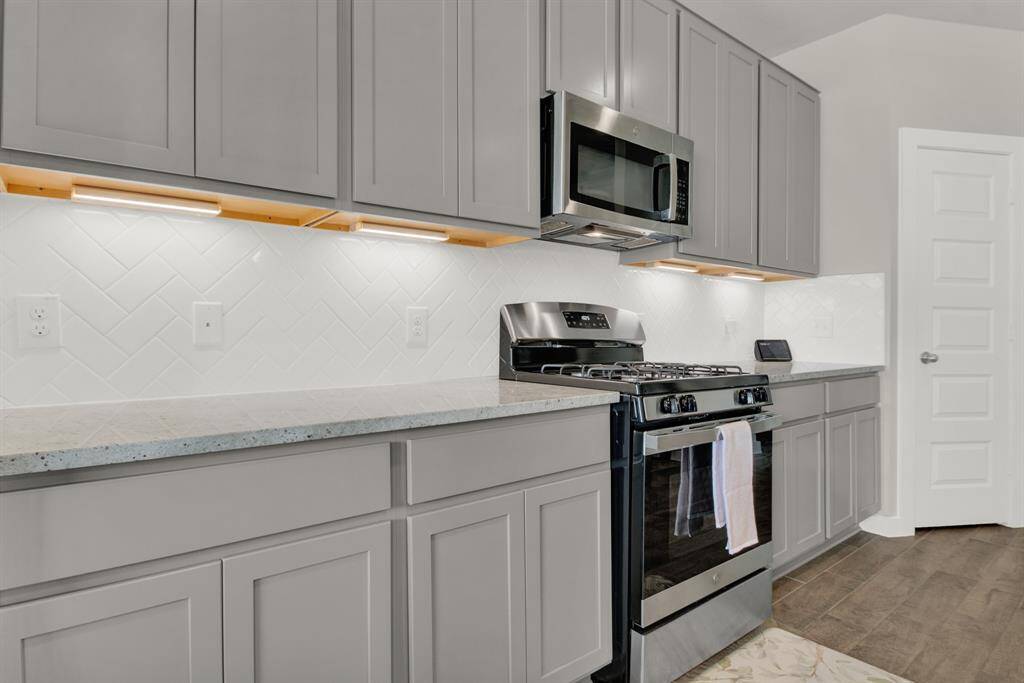 Modern kitchen with sleek gray cabinetry, stainless steel appliances, and a gas stove. Features a white herringbone tile backsplash and under-cabinet lighting.