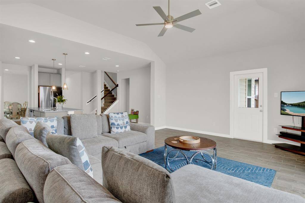Spacious open-concept living area with modern gray tones, sleek ceiling fan, and a view into the kitchen with stainless steel appliances. Natural light enhances the airy feel, and there's convenient access to an outdoor space.
