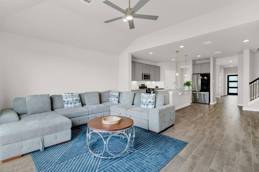 This spacious, open-concept living area leads into a modern kitchen with stainless steel appliances, pendant lighting, and a breakfast bar. The room is well-lit with recessed lighting and a ceiling fan, offering a contemporary and inviting atmosphere.