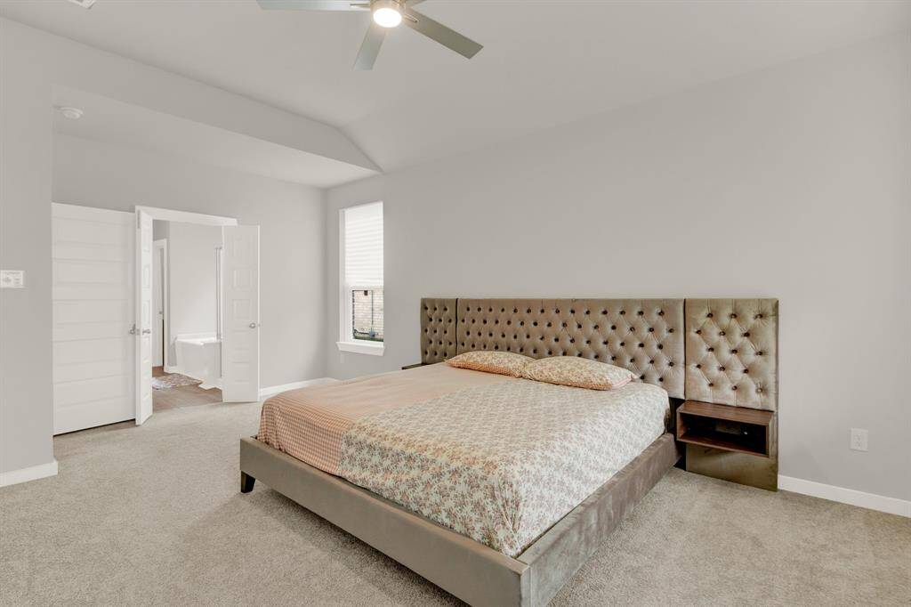 A spacious primary bedroom with neutral tones. The room has plush carpeting and two windows allowing natural light, offering a view of a serene pond outside.