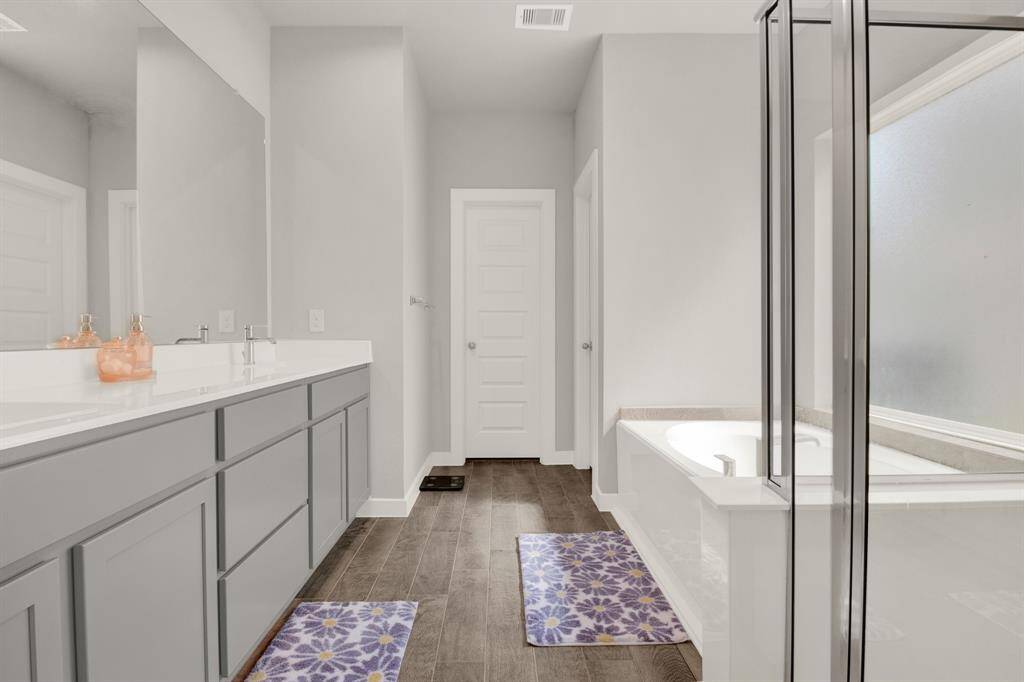 Modern bathroom featuring a double vanity with gray cabinets, a spacious shower, and a separate bathtub. The room is bright with neutral tones and elegant fixtures, complemented by wood-style flooring and decorative rugs.