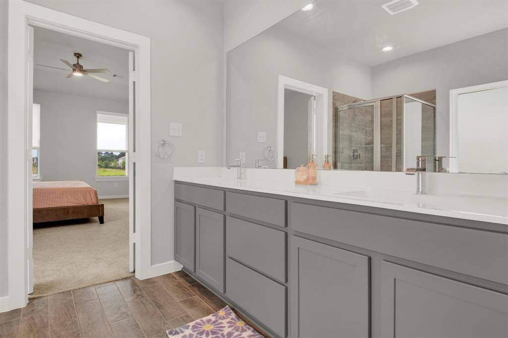 Modern bathroom with dual sinks and gray cabinetry. It features a large mirror and a glass-enclosed shower. The space leads into the primary bedroom with carpeted flooring and a ceiling fan.