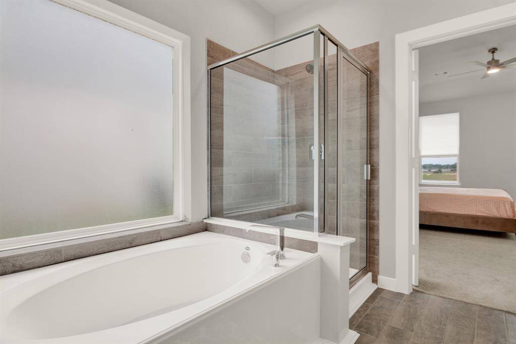 Modern bathroom with dual sinks and gray cabinetry. It features a large mirror and a glass-enclosed shower. The space leads into the primary bedroom with carpeted flooring and a ceiling fan.