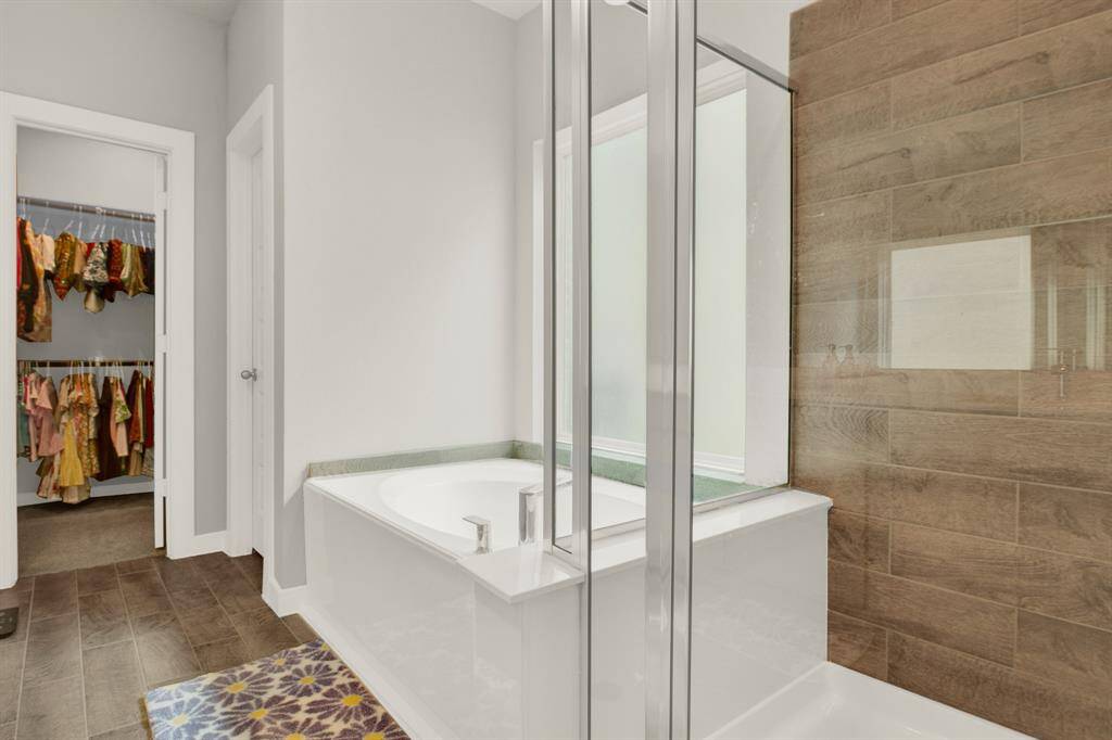 This bathroom features a modern design with a glass-enclosed shower and a separate bathtub. It has a wood-tiled accent wall and a doorway leading to a walk-in closet. The space is bright and functional, ideal for a comfortable lifestyle.