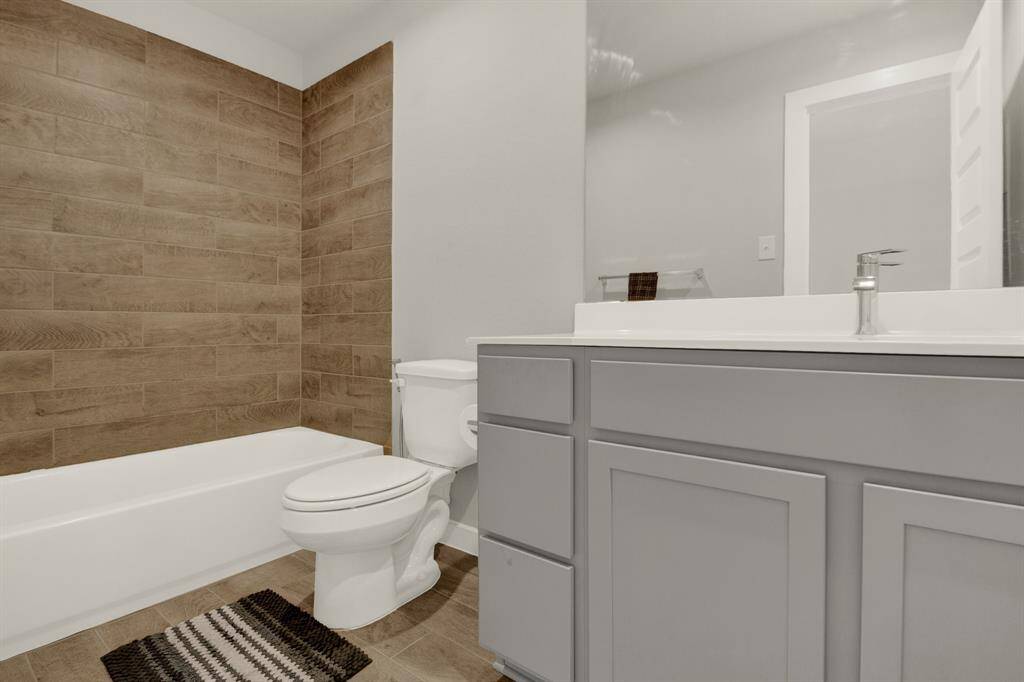 This secondary bathroom features a modern design with a sleek gray vanity, white countertop, and a large mirror. It has a bathtub with a wood-textured tile wall and coordinating floor tiles, creating a warm and inviting atmosphere.