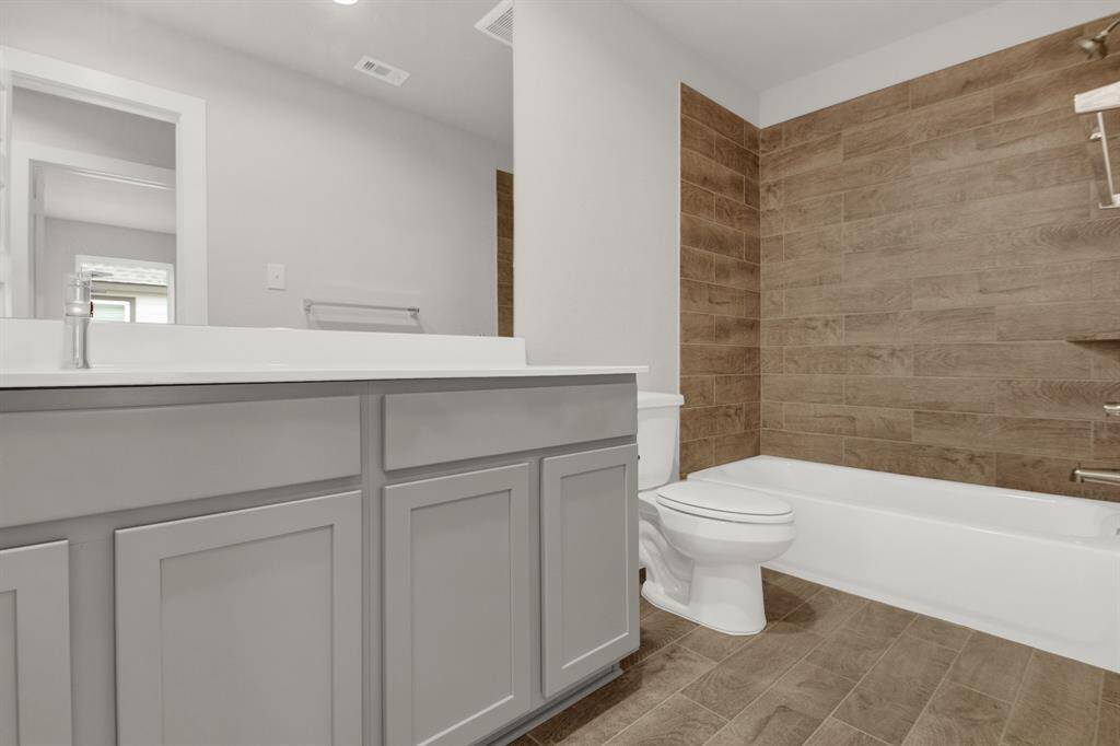 This secondary bathroom features a modern design with a sleek gray vanity, white countertop, and a large mirror. It has a bathtub with a wood-textured tile wall and coordinating floor tiles, creating a warm and inviting atmosphere.