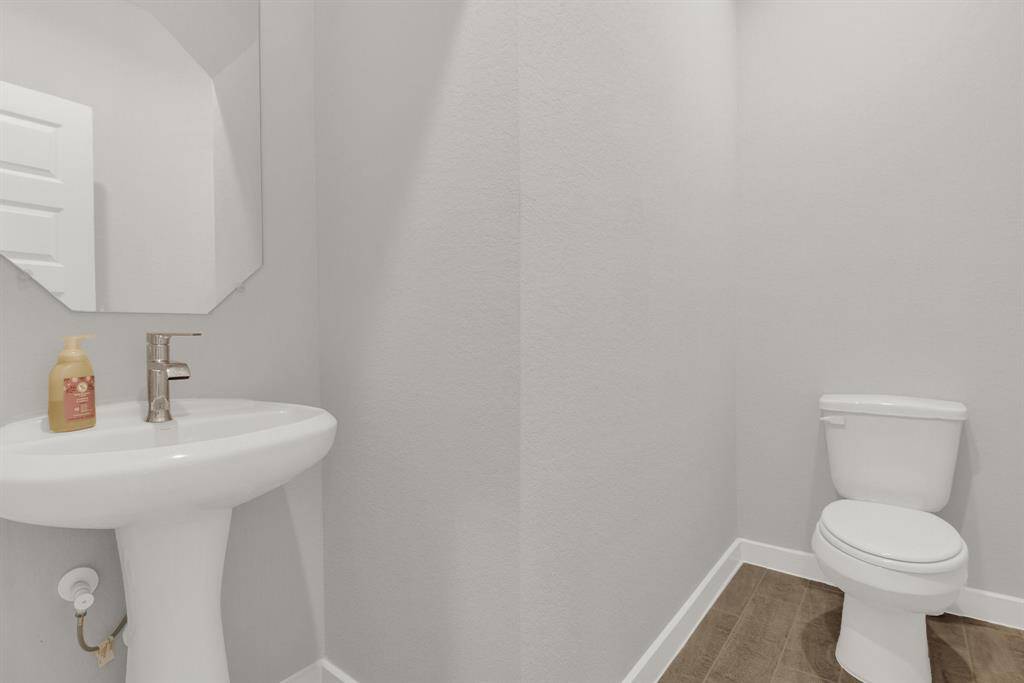 Modern half-bathroom with light gray walls, featuring a pedestal sink with a mirror above it and a toilet, complemented by a brown tiled floor.
