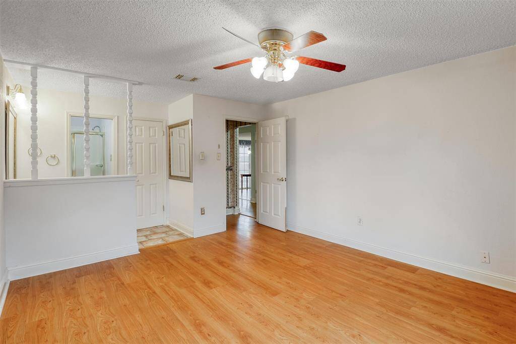 This primary bedroom features laminate wood flooring, white walls, and a ceiling fan with light. It includes a small partition with decorative spindles, leading to a bathroom with a visible shower. The space is bright and open, ideal for personalization.