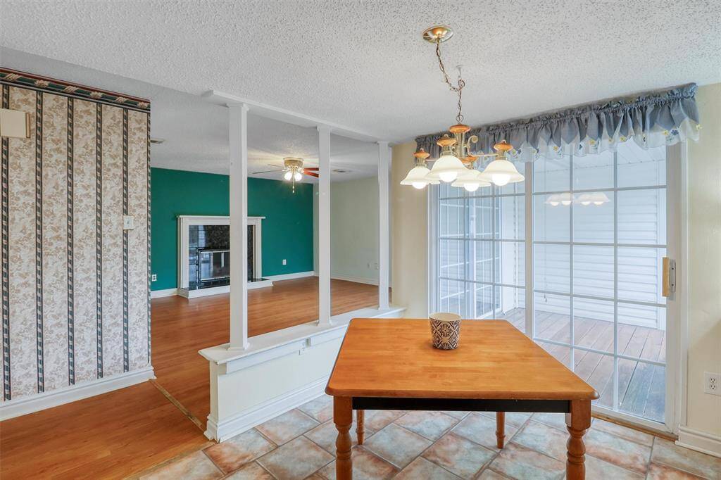 This photo showcases a bright kitchen area with vinyl flooring and a decorative light fixture. Adjacent is a living space with laminate wood floors, a ceiling fan, and a cozy wood burning fireplace, accented by a teal feature wall and patterned wallpaper.