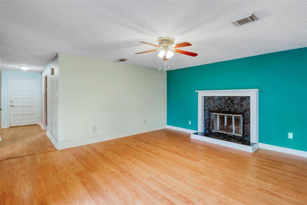 Bright family living area with laminate wood flooring, featuring a ceiling fan and a striking teal accent wall. The room includes a wood burning fireplace with a decorative mantle, creating a cozy focal point. Ample natural light enhances the spacious feel.