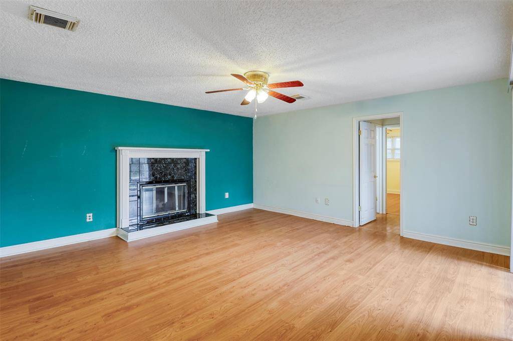 The room features laminate wood flooring, a teal accent wall with a wood burning fireplace, and a ceiling fan. It has ample natural light and leads to an adjoining room.