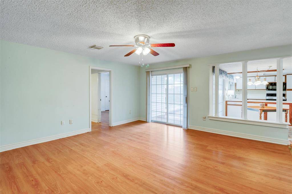 This room features laminate wood flooring and light walls, creating a bright and open feel. It includes a ceiling fan with a light, a sliding glass door leading to an outdoor area, and a windowed opening that connects to the kitchen, allowing for ample natural light and an airy atmosphere.