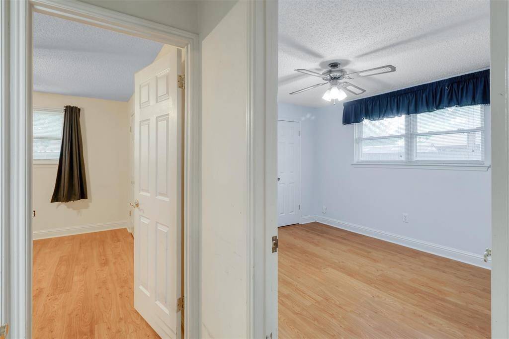 This photo shows a bright, dual-room view with laminate wood flooring. The main room features a ceiling fan and large windows with blue curtains, providing ample natural light. The secondary room is visible through an open doorway, offering additional space.
