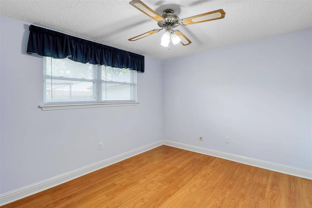 This room features light blue walls, laminate wood flooring, a ceiling fan with lights, and a window with a dark curtain valance, creating a bright and airy space.