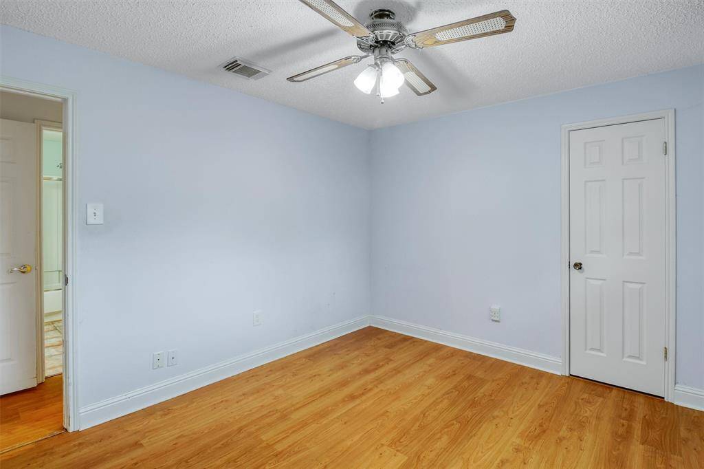 This room features light blue walls, laminate wood flooring, a ceiling fan with lights, and a door leading to an adjacent bathroom. Ideal for a bedroom or office space.