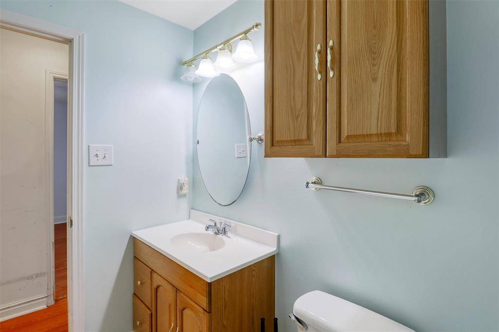 This is a compact bathroom featuring light blue walls, a wooden vanity with storage, an oval mirror, and overhead lighting. There’s a wall-mounted cabinet for additional storage, and a towel bar is present above the toilet.