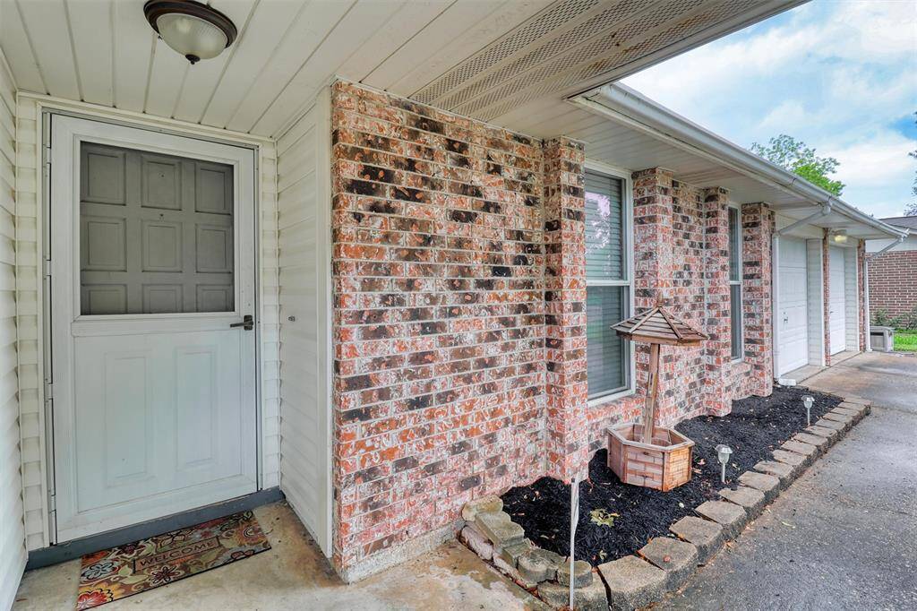 Charming front entrance with a brick facade and white siding, featuring a covered porch area. The entryway includes a welcoming front door and a decorative planter bed alongside.