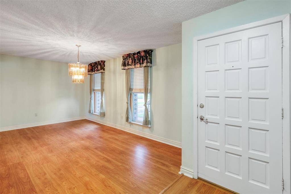 This photo shows a bright, open entryway featuring a white door with decorative panels, light laminate wood floors, and a stylish chandelier. There are two large windows with floral valances, providing plenty of natural light.