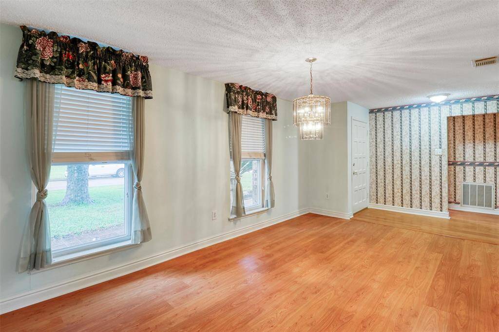 This dining or flex room features a cozy, well-lit space with laminate wood floors and two large windows adorned with floral curtains. The ceiling has a decorative light fixture, and there's an accent entry wall with patterned wallpaper.
