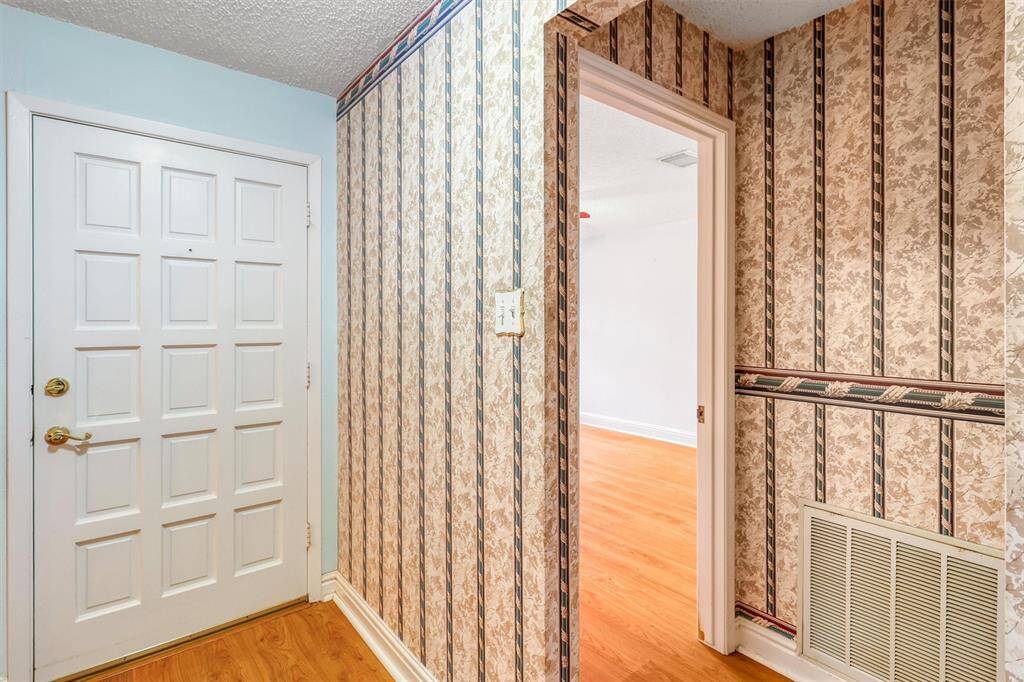 This entryway features a classic white door with paneled design and a gold handle. The walls are adorned with vintage patterned wallpaper, leading into a room with light laminate wood flooring.