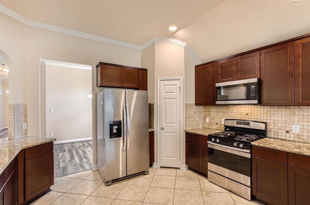 This kitchen features modern stainless steel appliances, including a fridge and gas stove, with dark wood cabinetry and granite countertops. The tile flooring and neutral backsplash add to its sleek design.