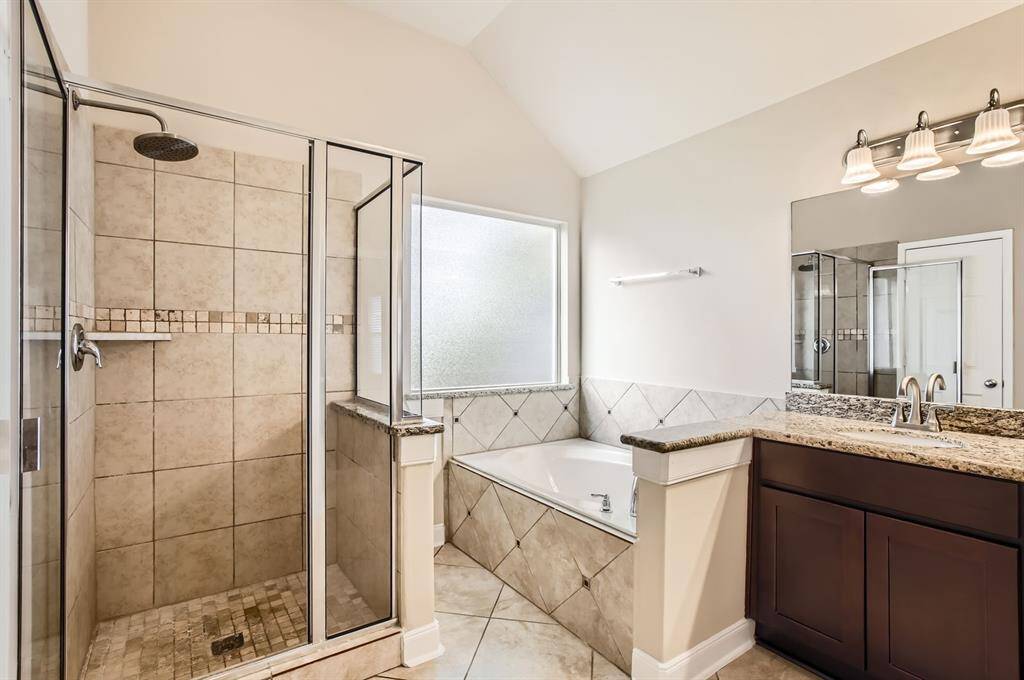 This primary bathroom ensuite features a spacious walk-in shower with glass doors and tile walls, a large soaking tub, and a granite-topped vanity with dual sinks. It has ample lighting and a frosted window for natural light.