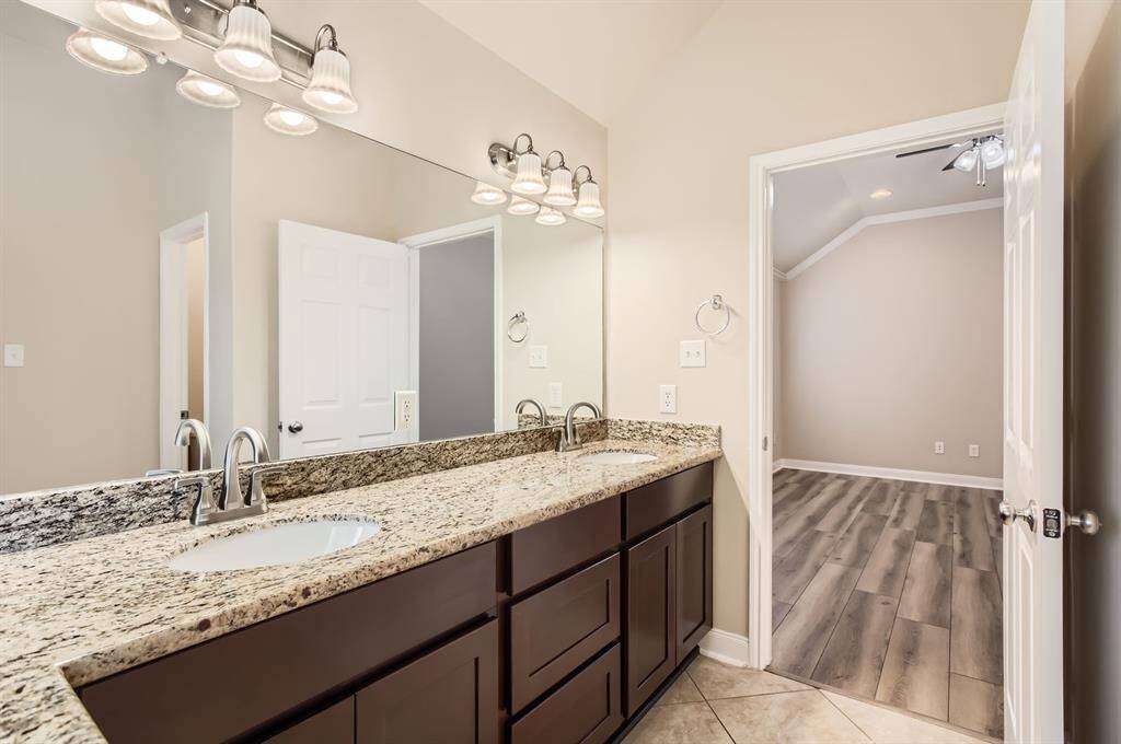 This primary bathroom with a granite double vanity, updated light fixtures, and a large mirror. The room has neutral tile flooring and opens into a primary bedroom, adding a contemporary feel.